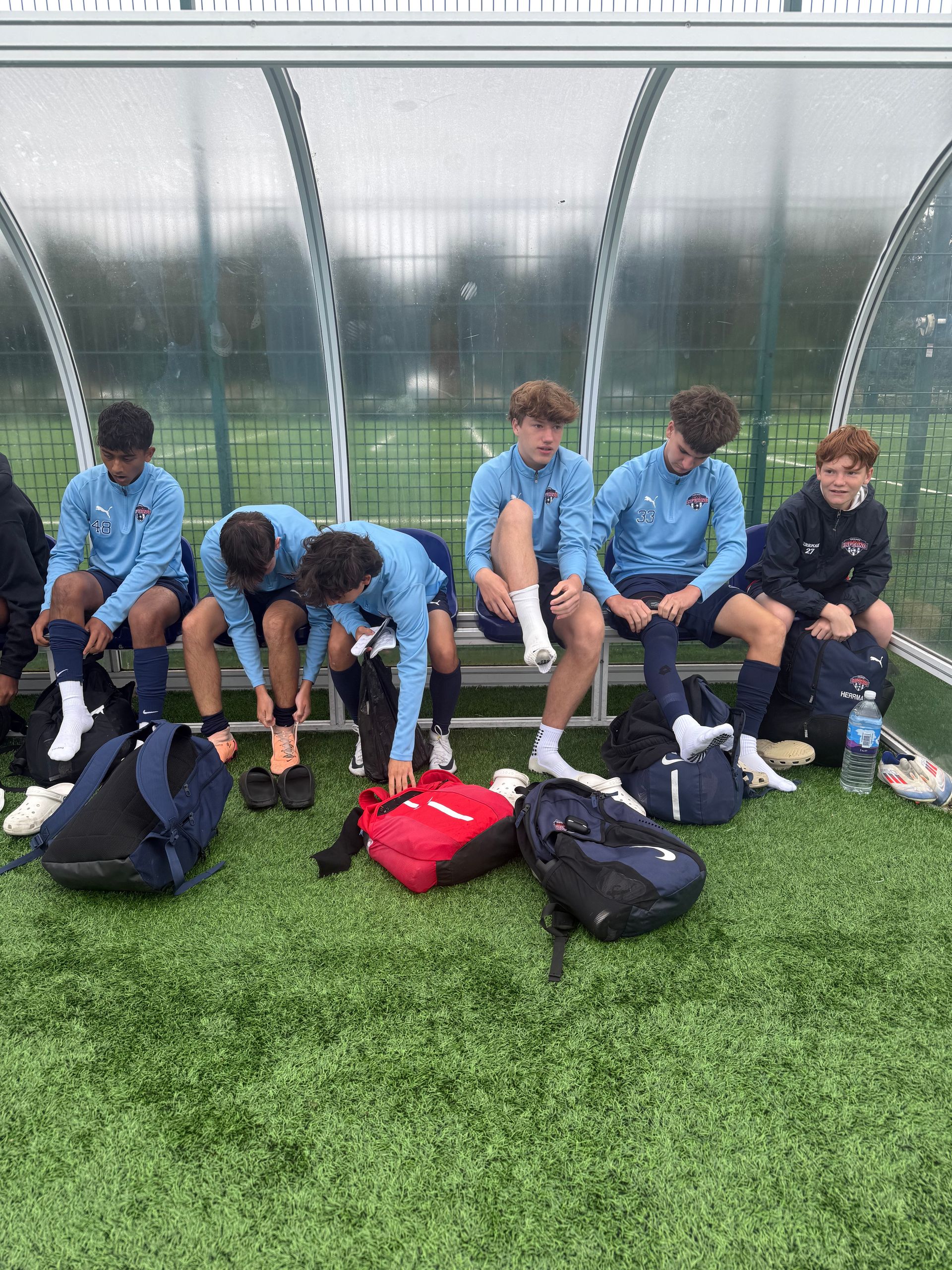 Six soccer players in light blue jerseys seated on a bench, preparing their gear on a field.