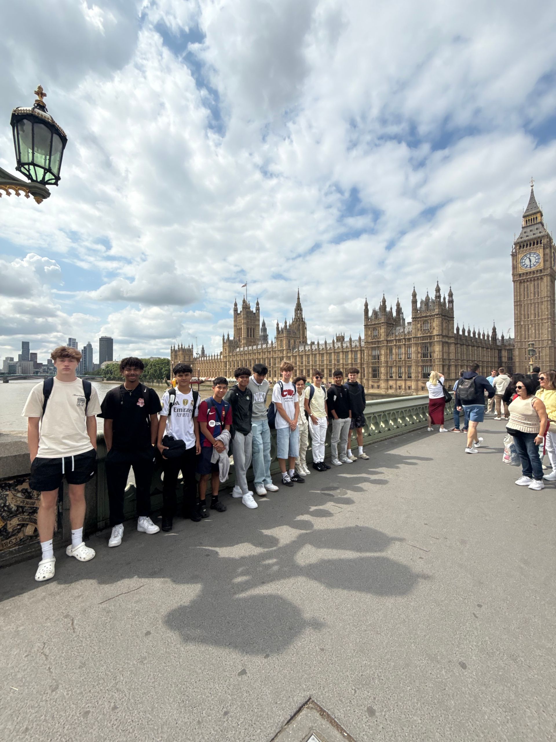 Group of people posing on a bridge in front of the Palace of Westminster and Big Ben in London, under a cloudy sky.