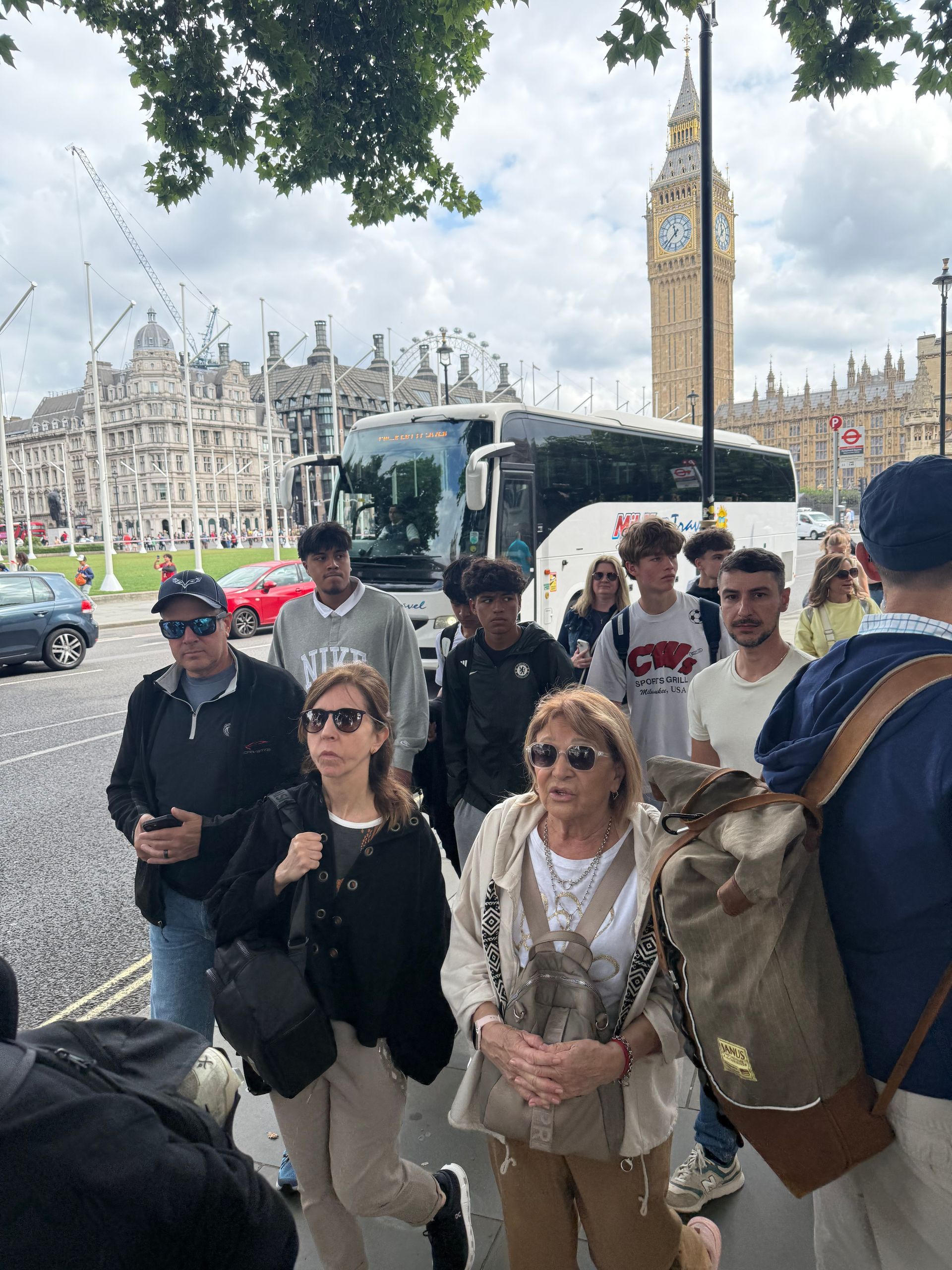 People stand on a sidewalk near Big Ben in London, a bus and other buildings in the background.