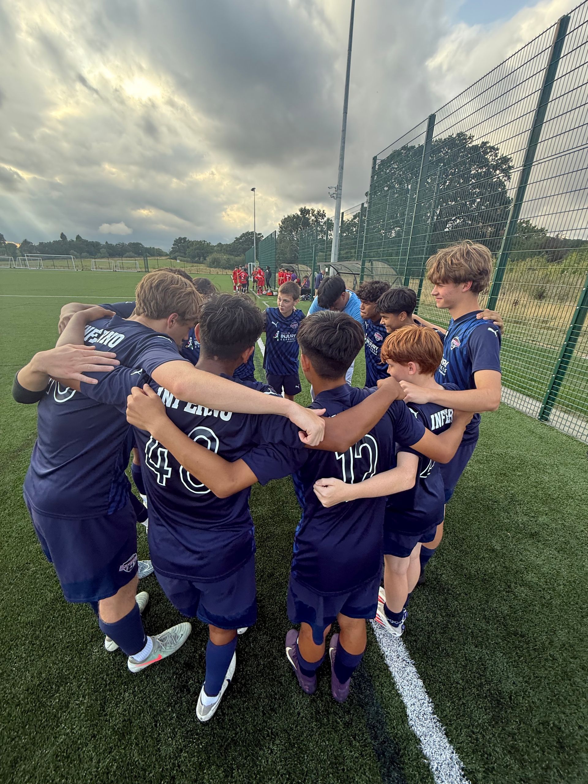 Soccer team huddled together on the field, wearing navy blue uniforms.