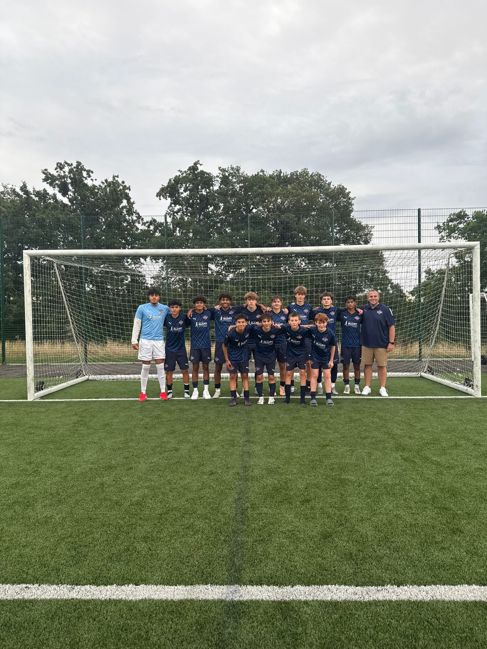 Soccer team poses in front of goal on green field, cloudy sky. Players in navy uniforms, coach in beige.