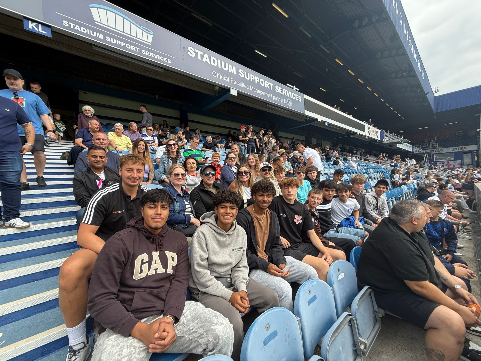 People in stadium seating, watching an event. Blue and white striped seats. Cloudy sky.