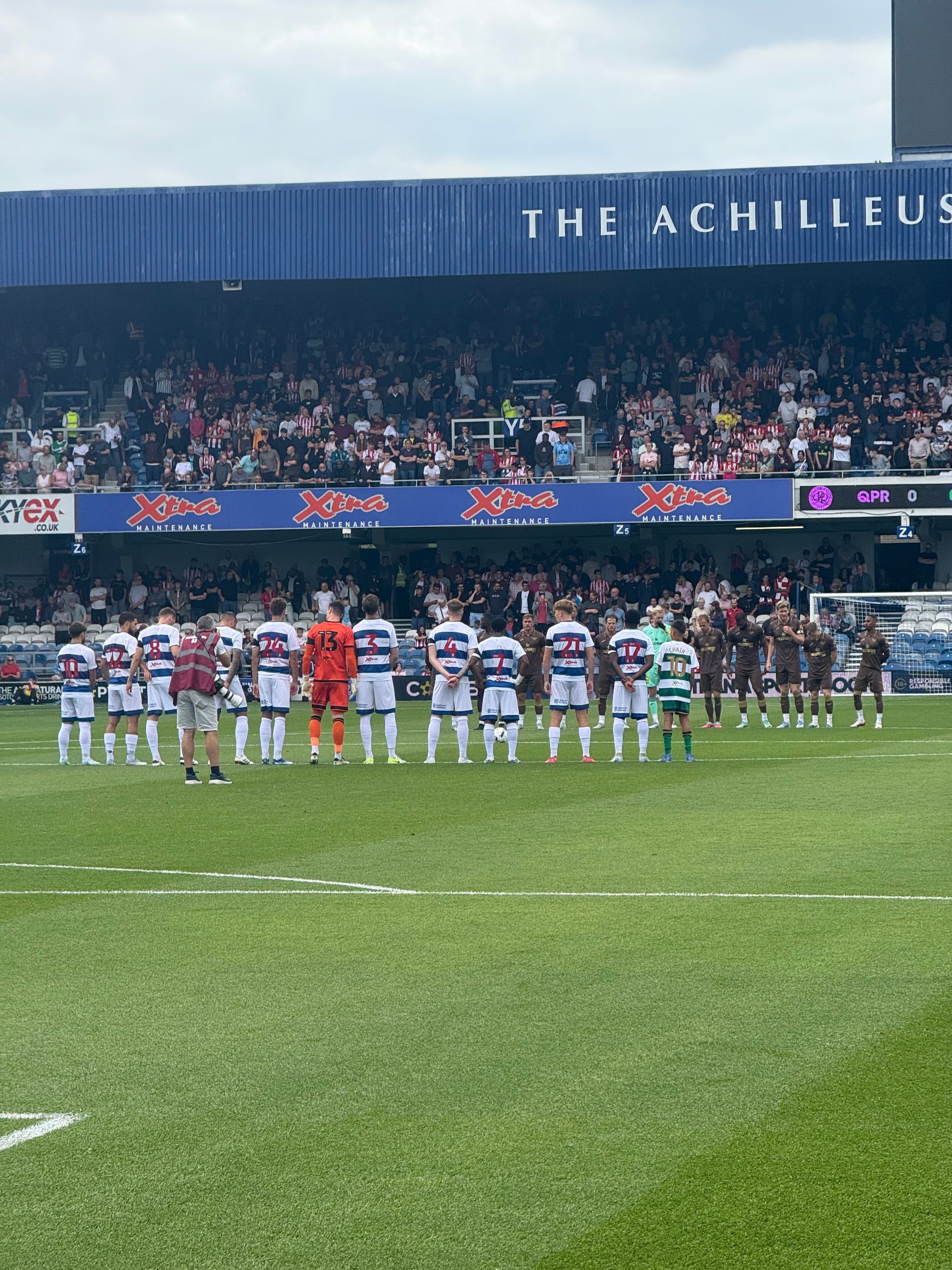 Soccer teams lined up on a green field at a stadium. Crowd in the background.