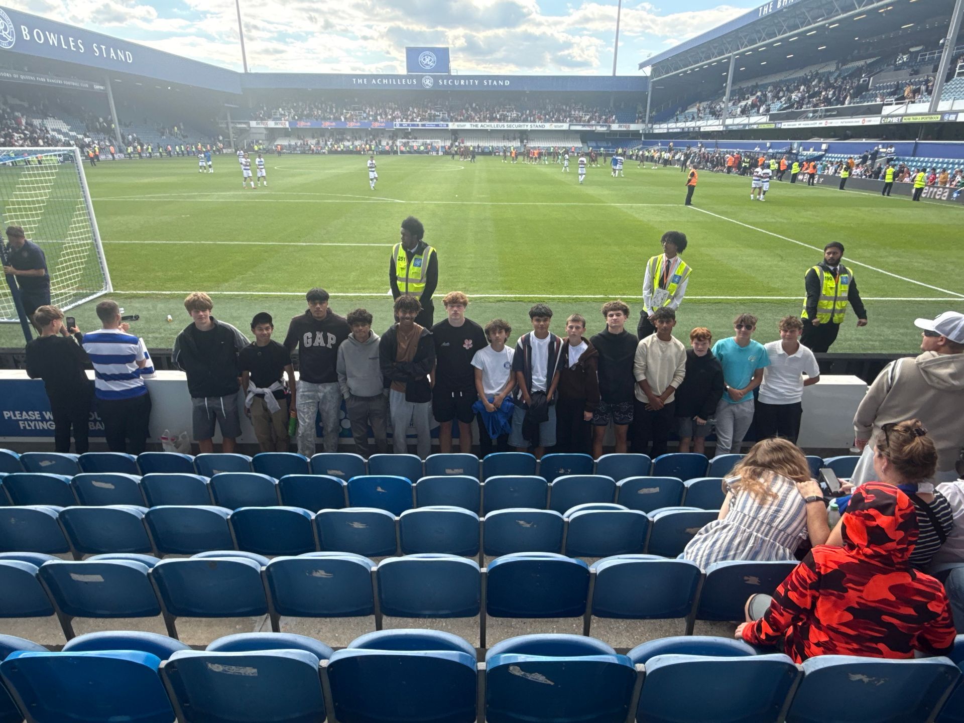 Group of people standing in front of a soccer field at a stadium. Several are wearing dark clothes.