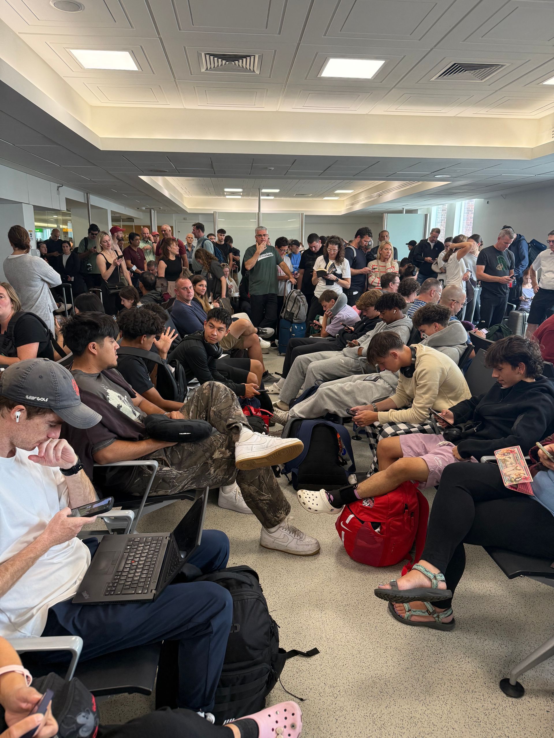 People sitting and standing in a crowded airport terminal. Some use phones or laptops. Luggage is scattered.