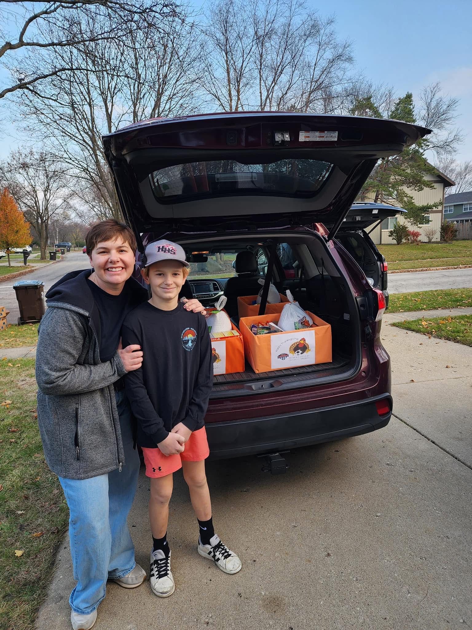 Woman and child stand near an open car trunk filled with boxes; autumn street setting.