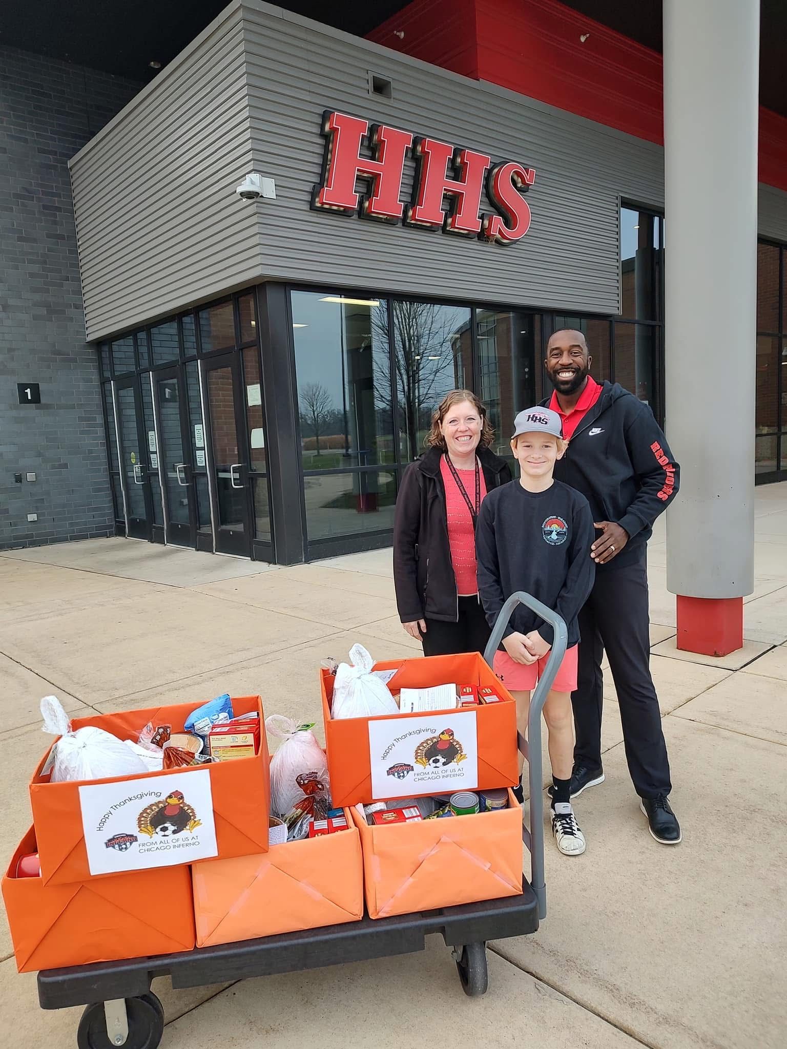 Three people stand with a cart of food boxes in front of HHS building.