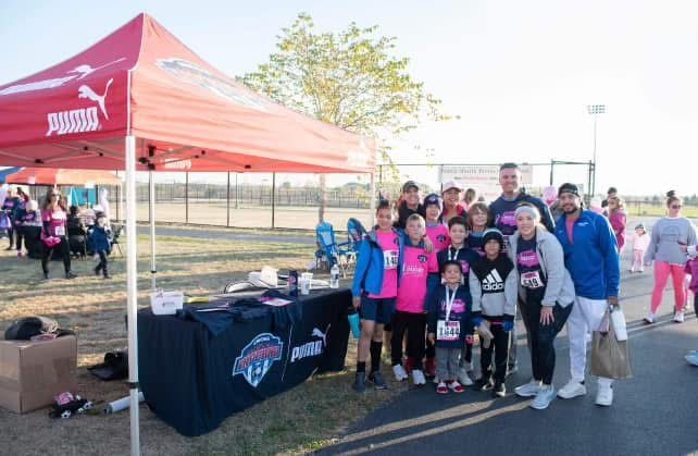 Group of people posing by a pink Puma tent at an outdoor event; some wearing pink and race bibs.