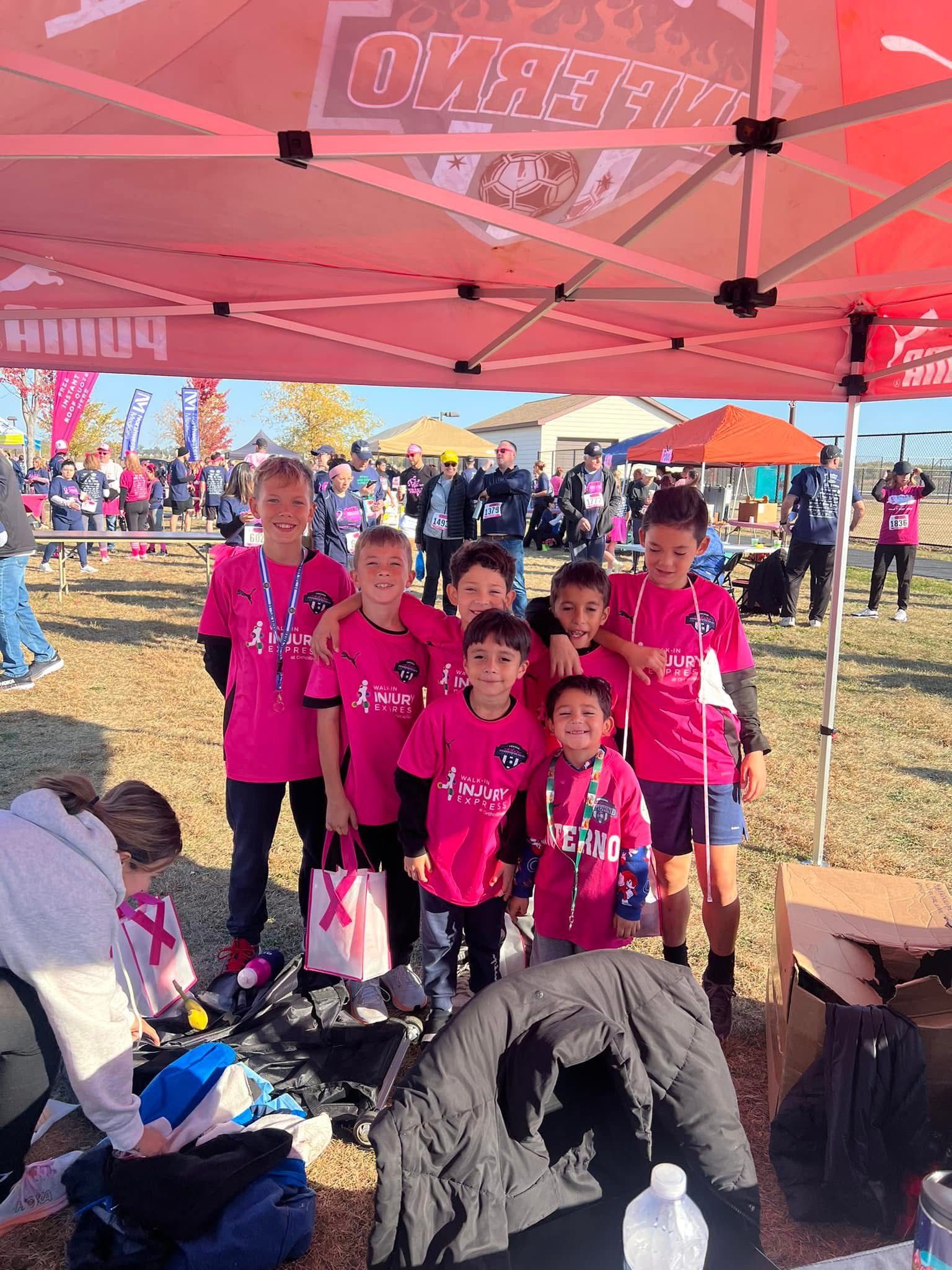 Soccer team in pink breast cancer awareness shirts under a canopy, posing for a photo at an outdoor event.
