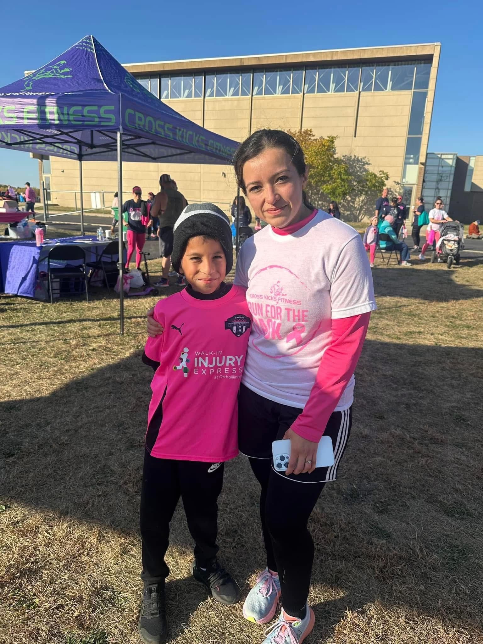 Woman and child in pink attire pose outdoors. Background shows a tent and a building.