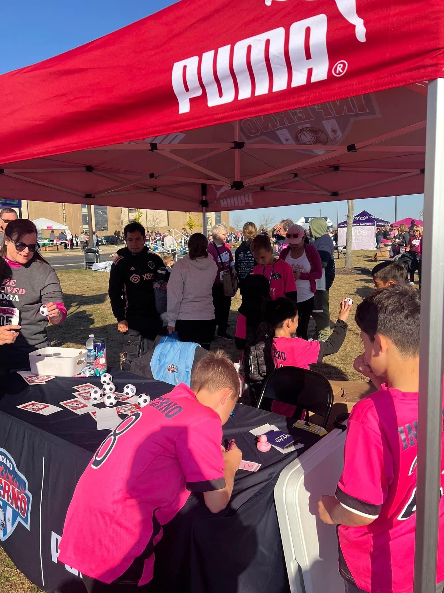 People gathered at a Puma tent, some wearing pink shirts. A table with items, sunny outdoor setting.