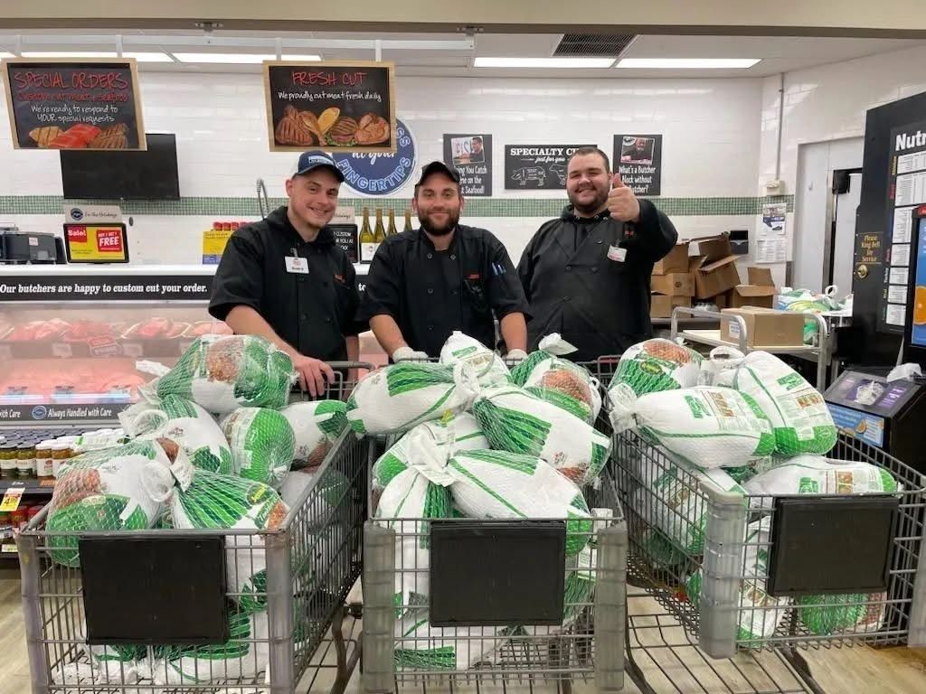 Three men in aprons stand by shopping carts overflowing with turkeys in a supermarket meat department.