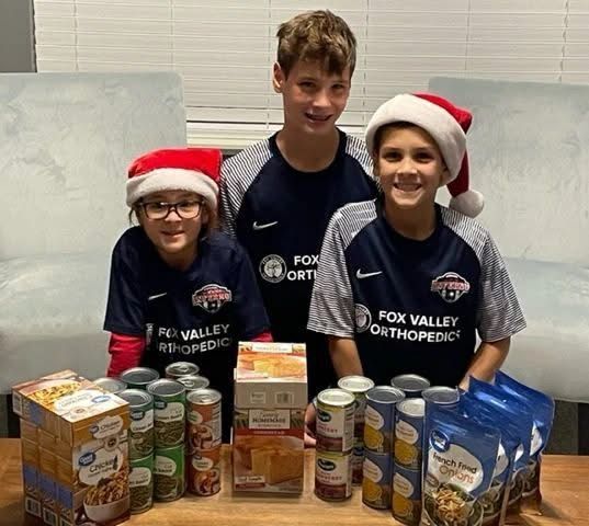 Three children in soccer shirts and Santa hats with donated food items.