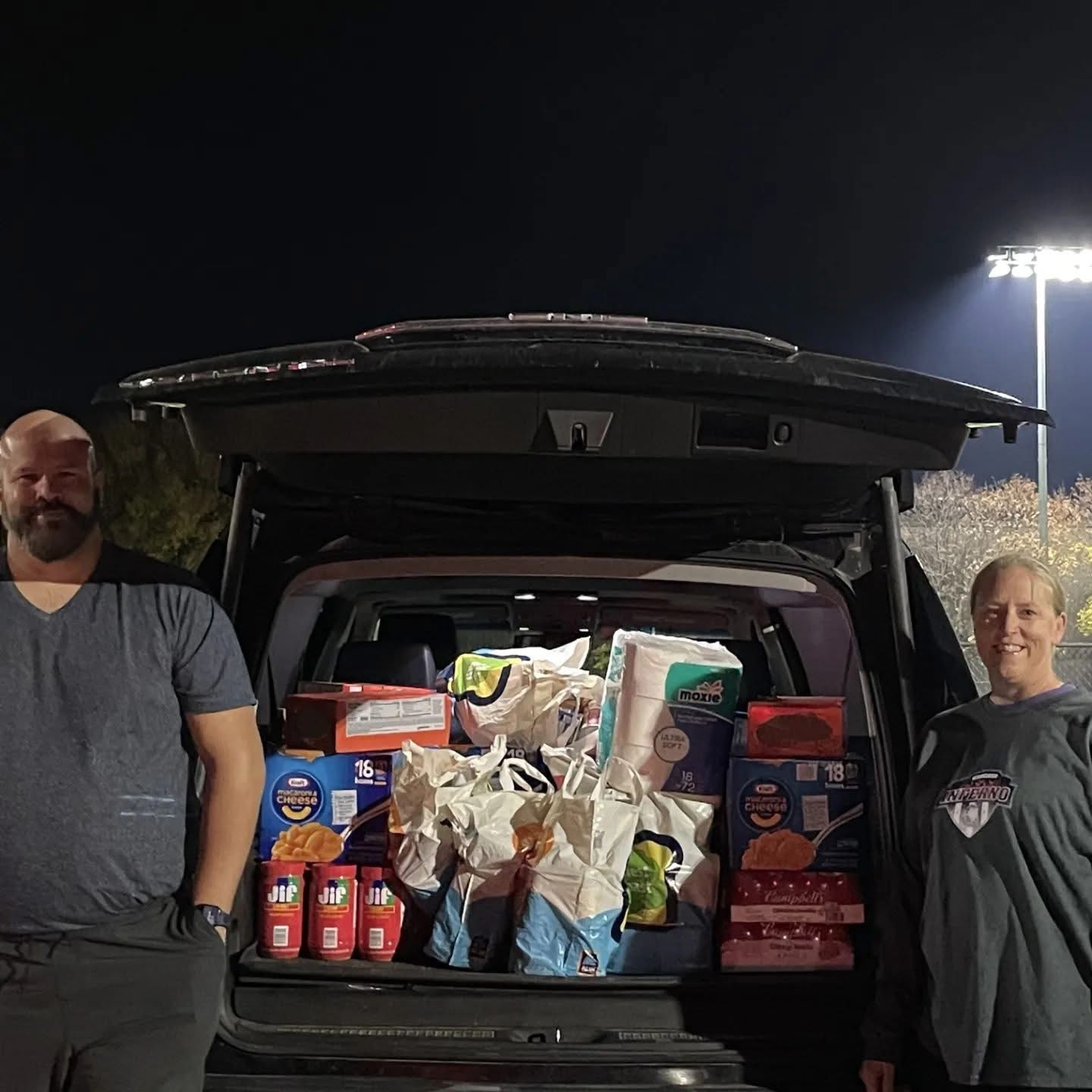 Two people stand near a car's open trunk filled with donated groceries and supplies at night.