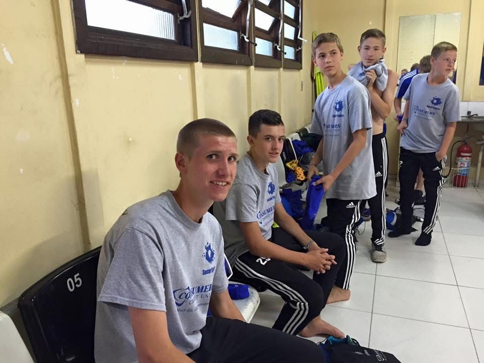 Group of young men in gray shirts, possibly a sports team, in a locker room, some smiling.