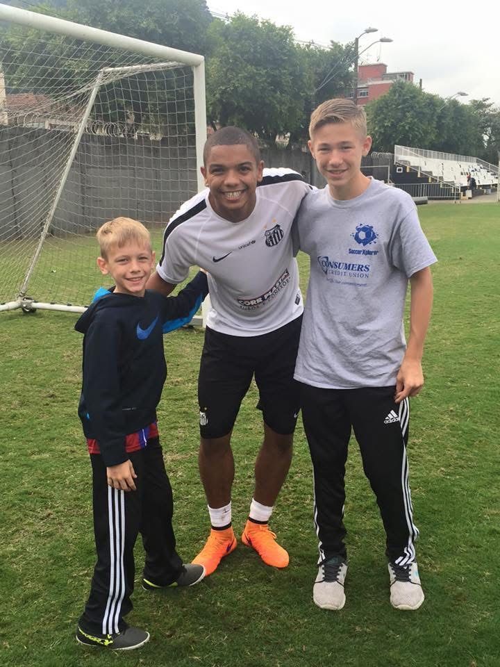 Three people standing on a soccer field. Man in white shirt has arms around two boys. They are smiling.
