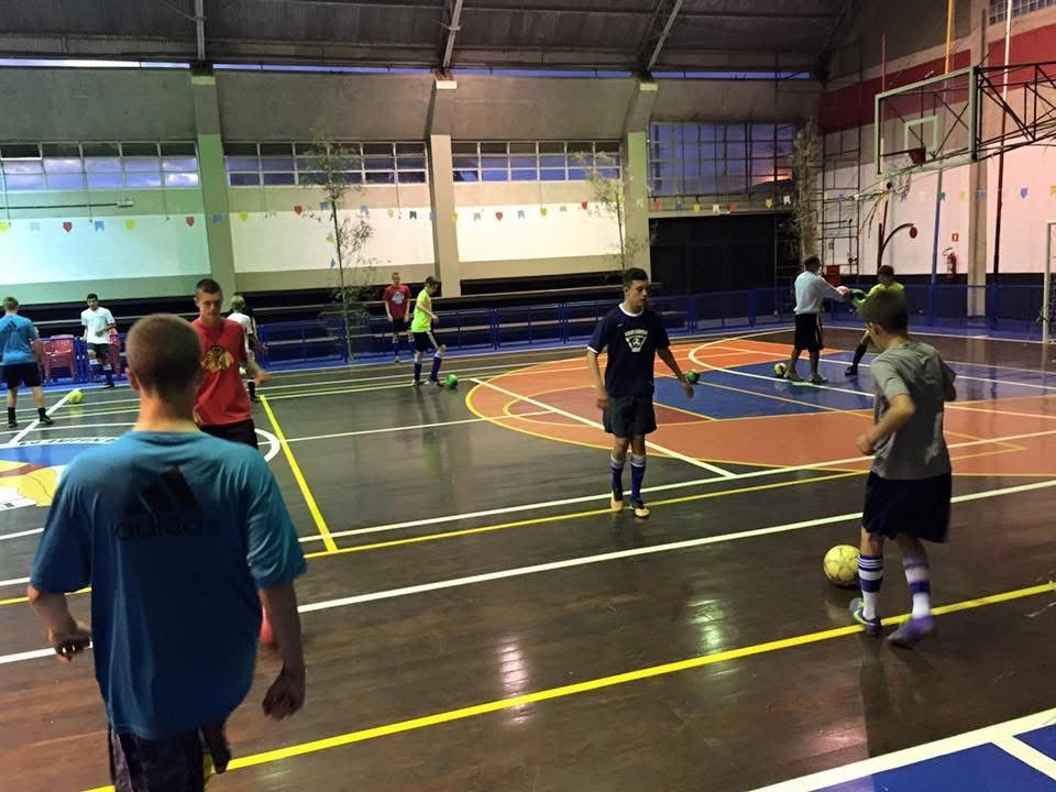 People playing soccer on an indoor court with dark wood floors and painted lines.