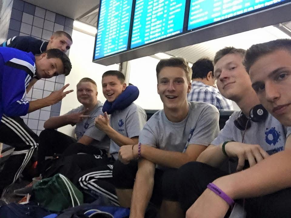 Group of young men in a public space, smiling and posing for a photo. Airport setting with flight information display.