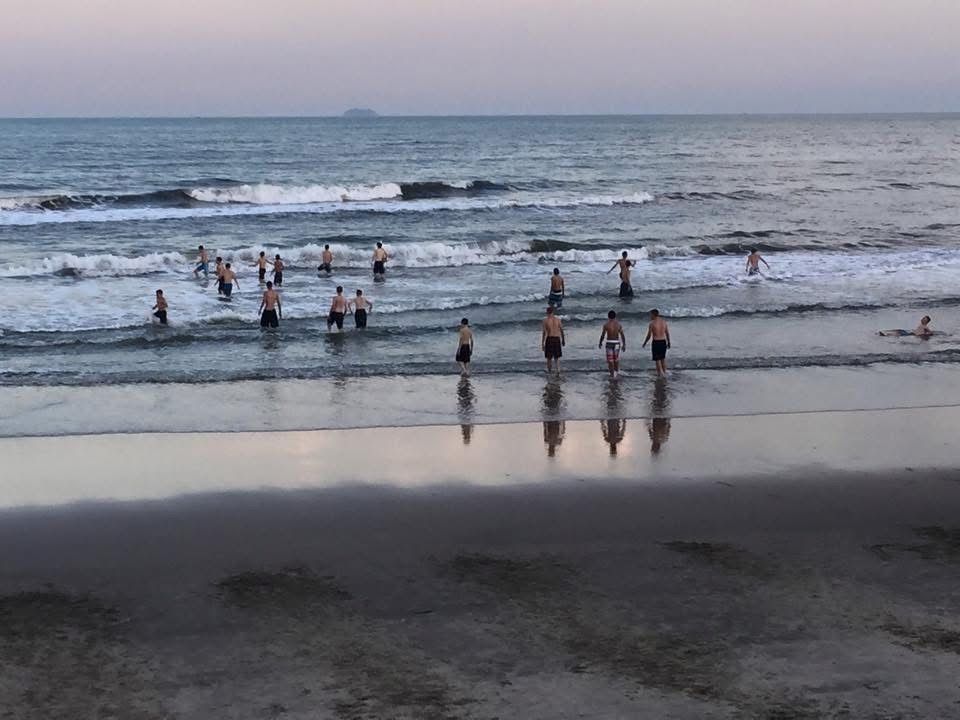 People wading and swimming in the ocean at sunset. The beach reflects the calm water and figures.