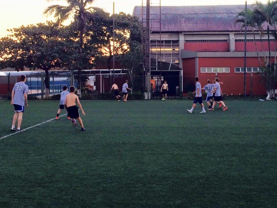 People playing soccer on a green field in front of a building. Sunset in the background.
