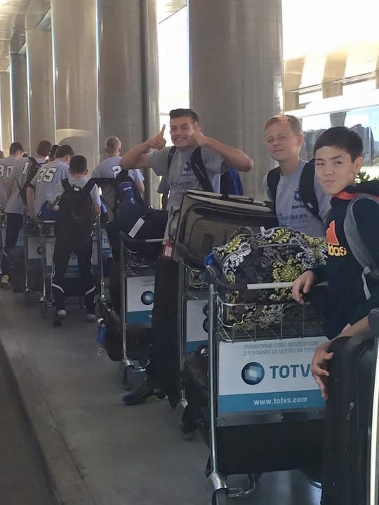 People with luggage on airport carts, smiling and giving thumbs-up. Outdoor setting with pillars, possibly boarding a bus.