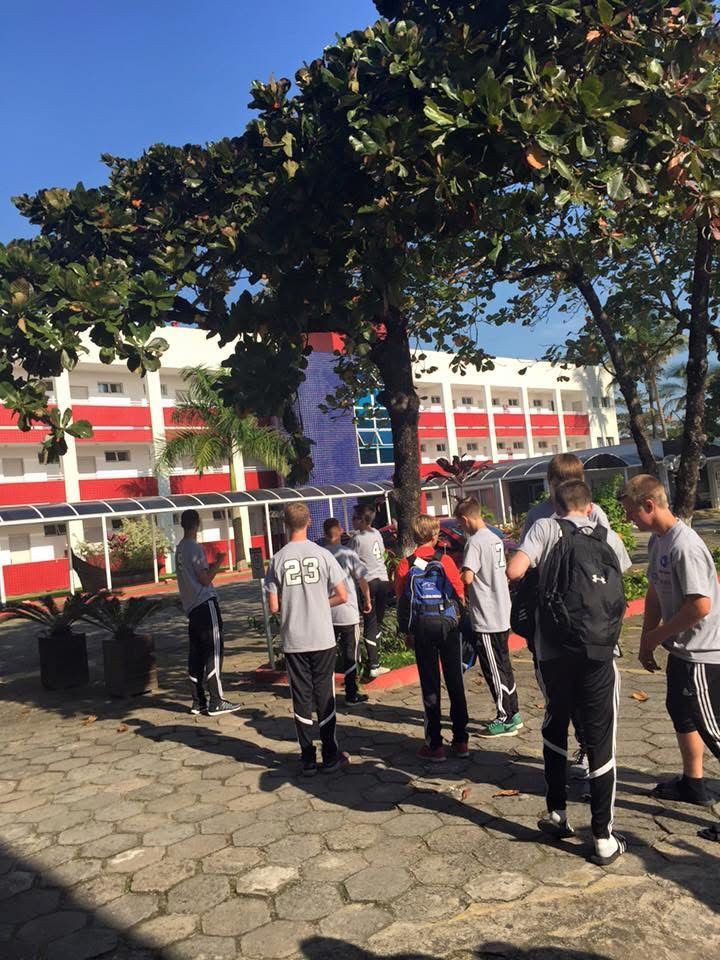Group of students outside a red and white building, standing under a tree on a sunny day.