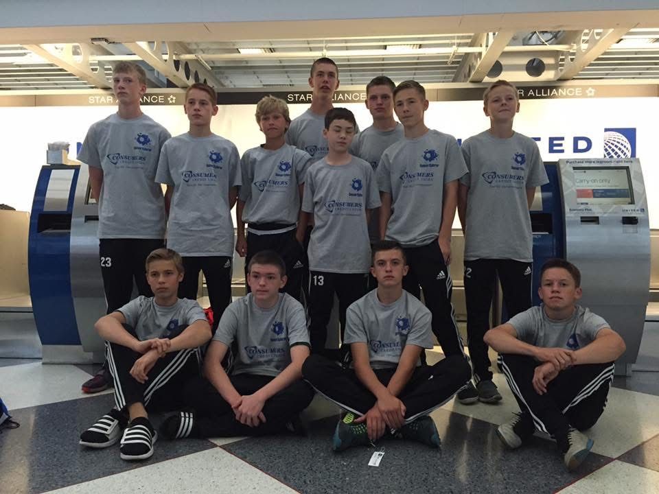 Soccer team posing together in front of United Airlines kiosks at an airport. They wear matching gray shirts and black pants.