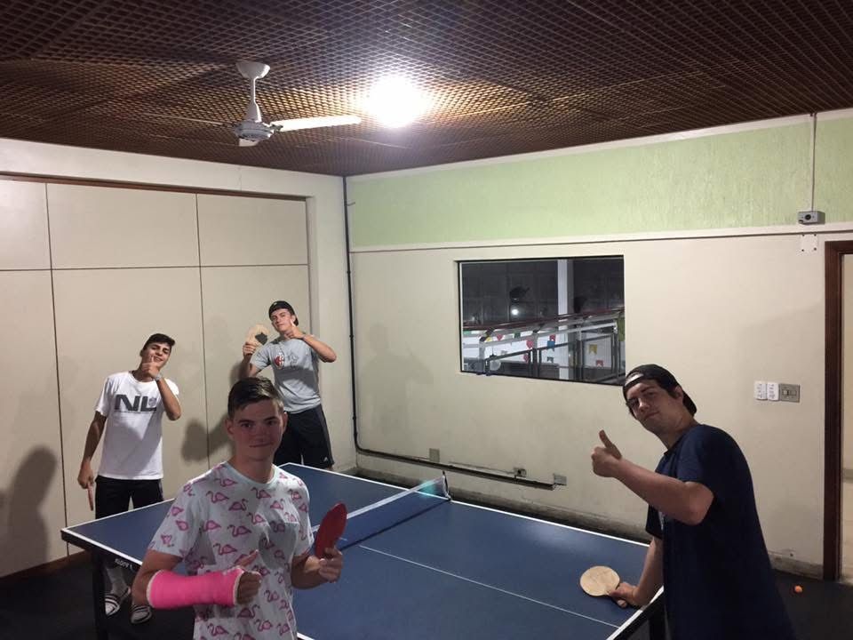 Four people playing ping pong in a room, two holding paddles, one with an arm cast, smiling.