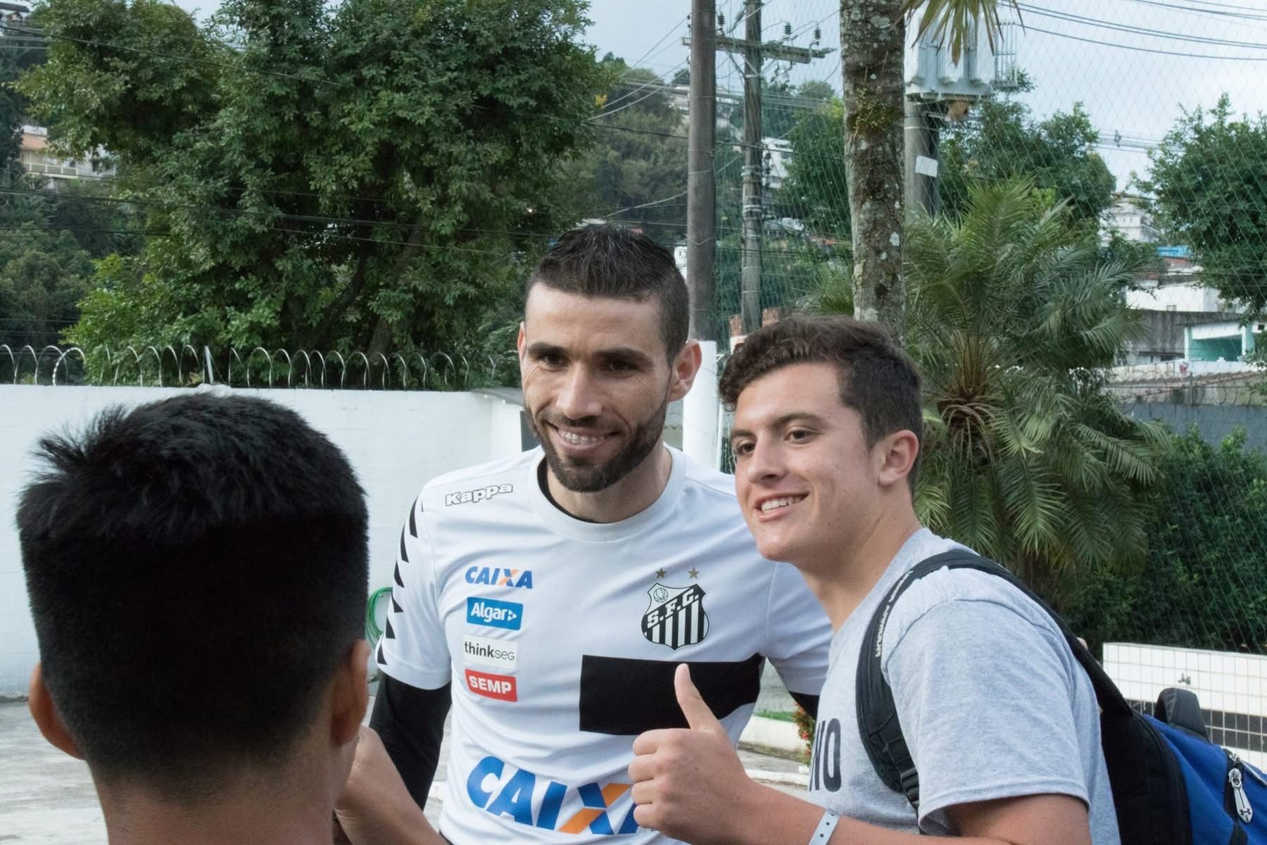 Soccer player poses with fan, thumbs up. Both are smiling outdoors. Player wears team jersey.