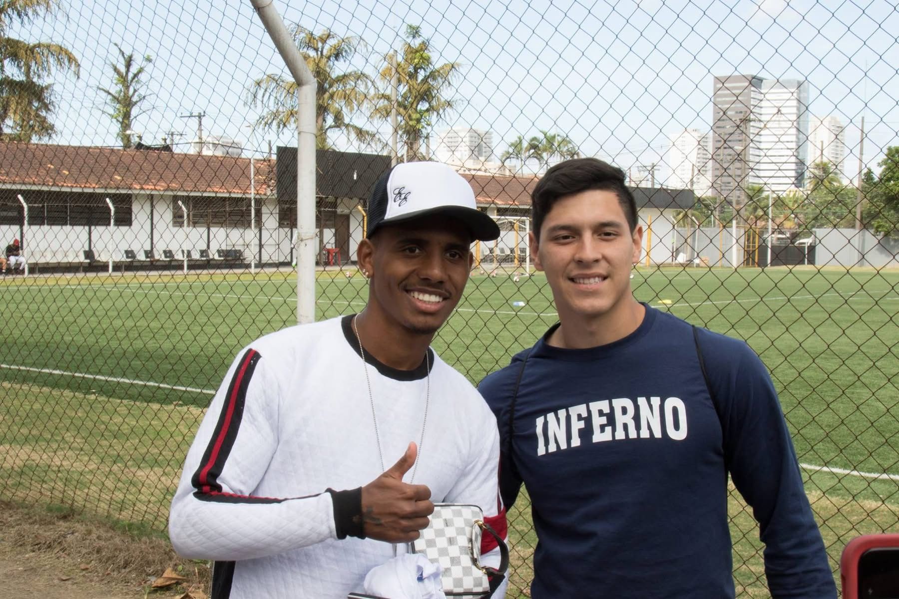 Two men smile, posing outdoors near a sports field. One gives a thumbs-up and wears a hat.
