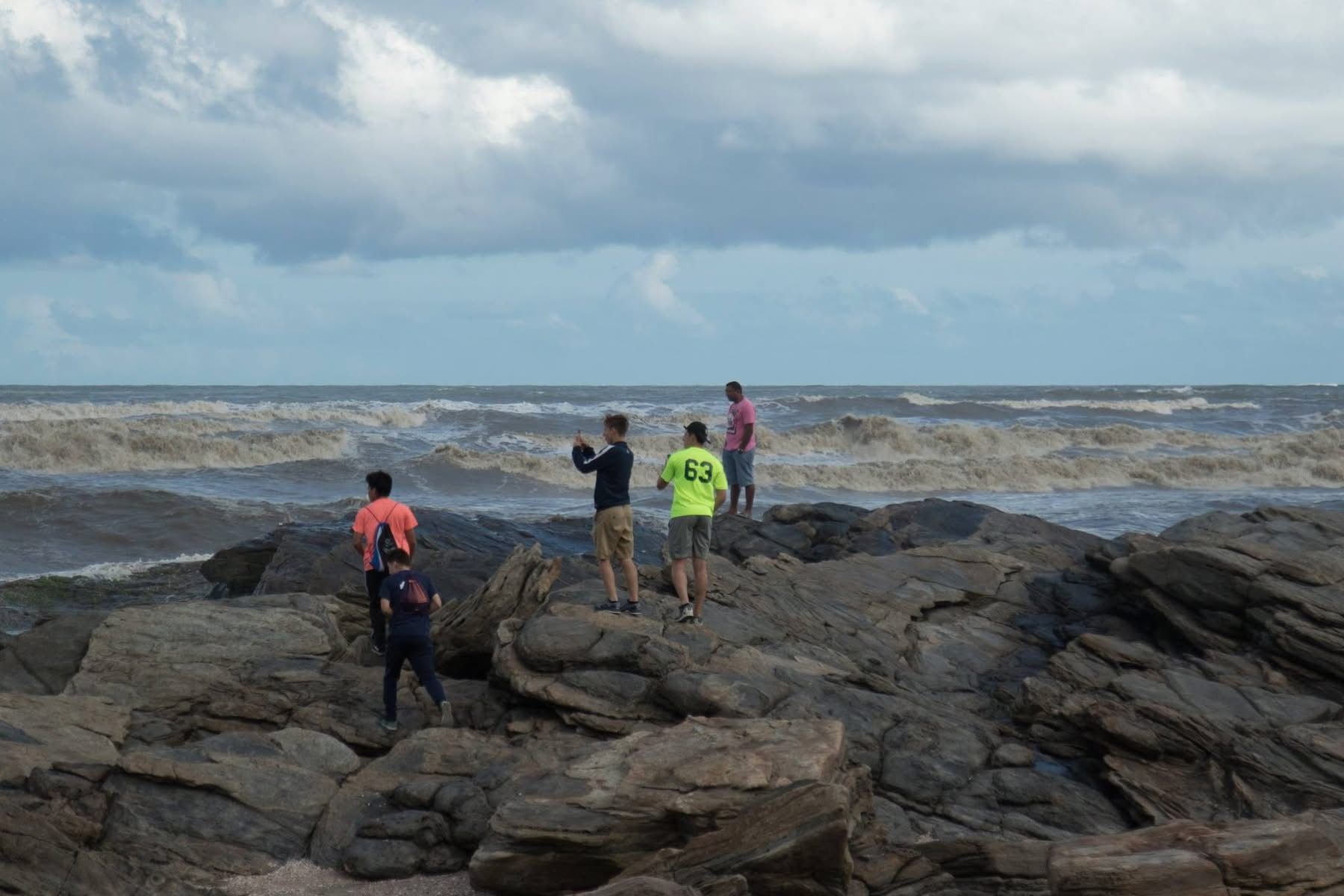 People on rocky shore looking at choppy ocean under cloudy sky.