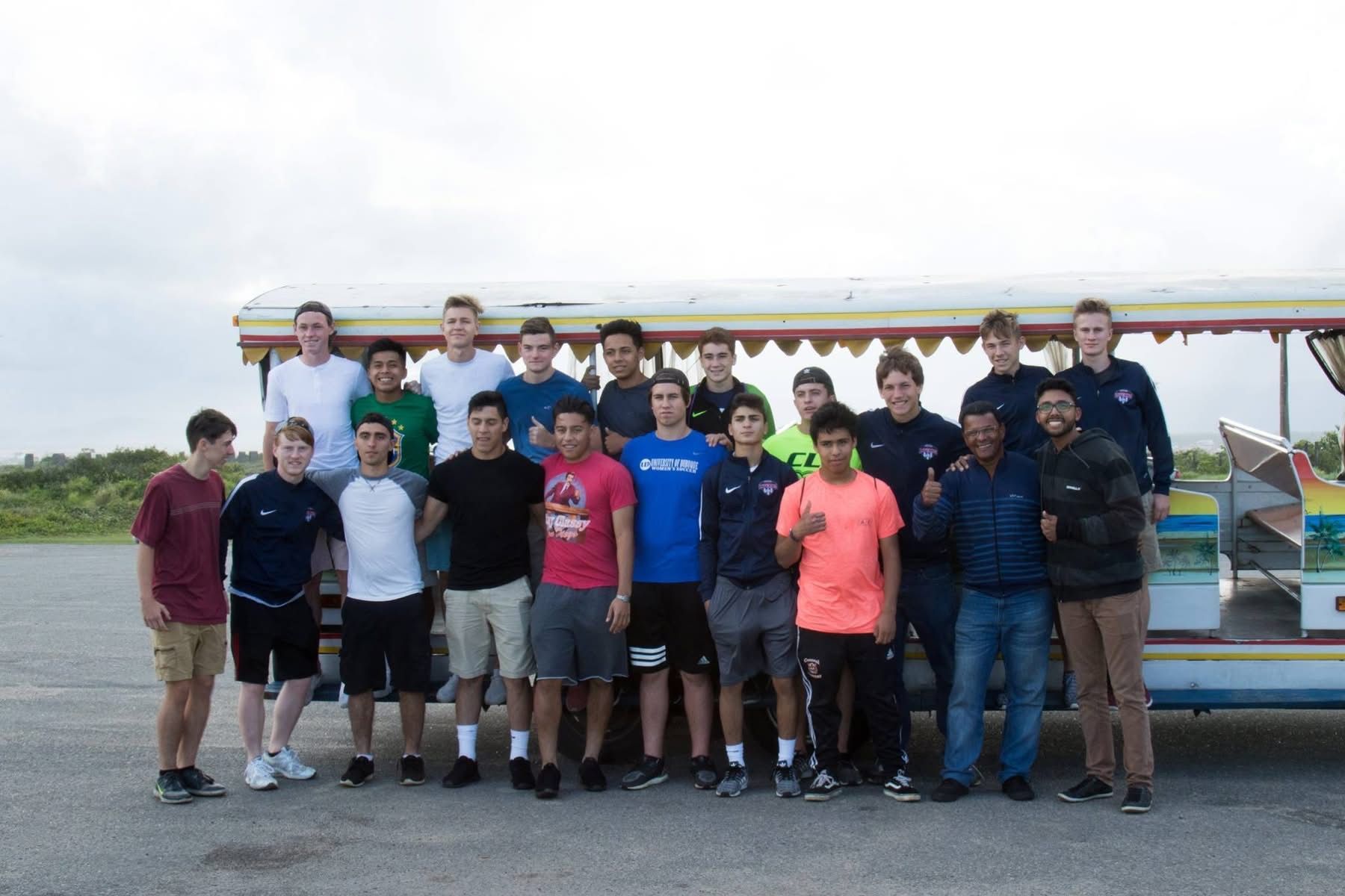 Group of young men stand in front of a colorful vehicle. Cloudy sky. Some are wearing sports attire.