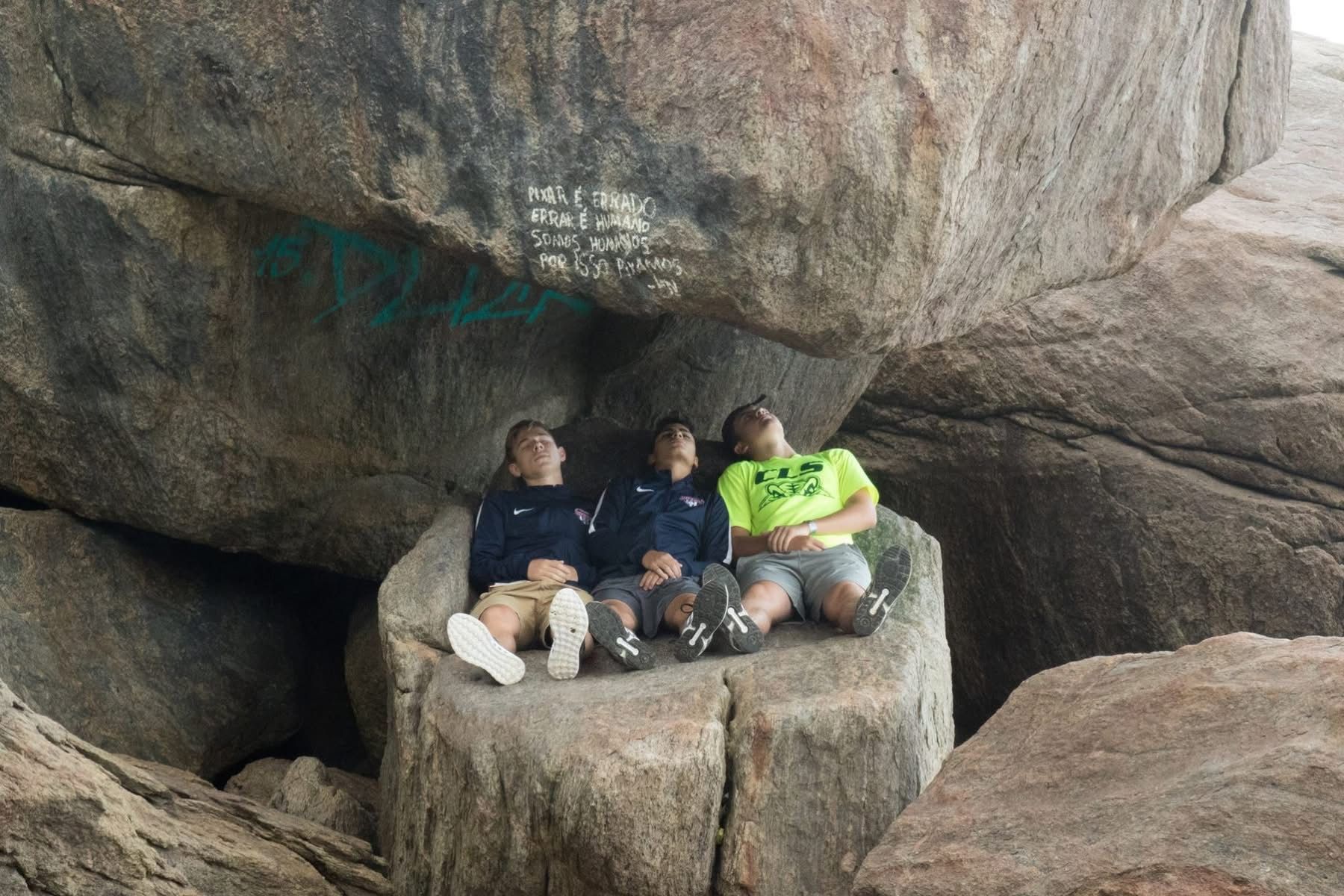 Three people sitting on a large rock under larger rocks, looking upward.