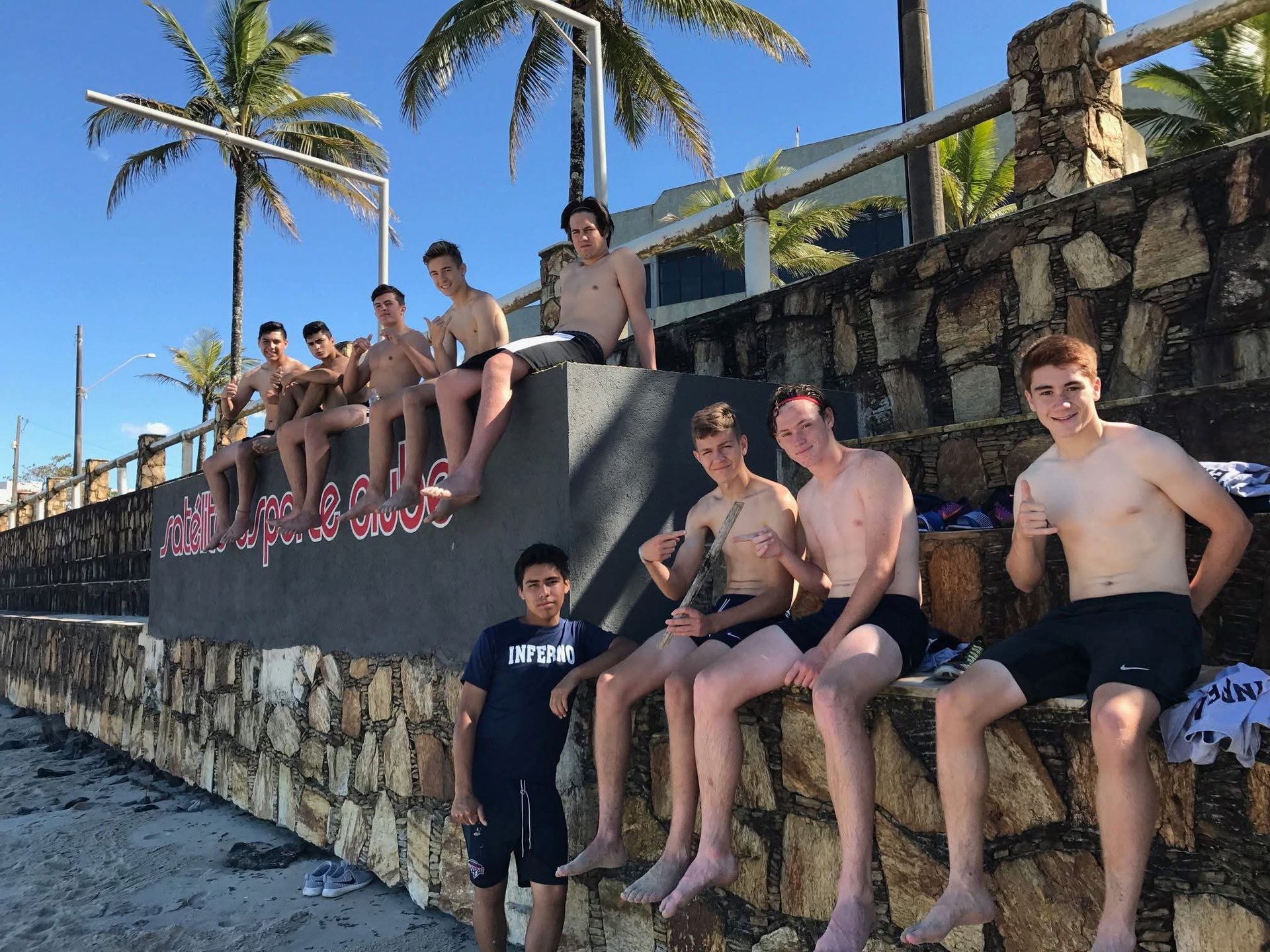 Group of shirtless people sitting on a wall near the beach. Some are smiling, others are looking at the camera.