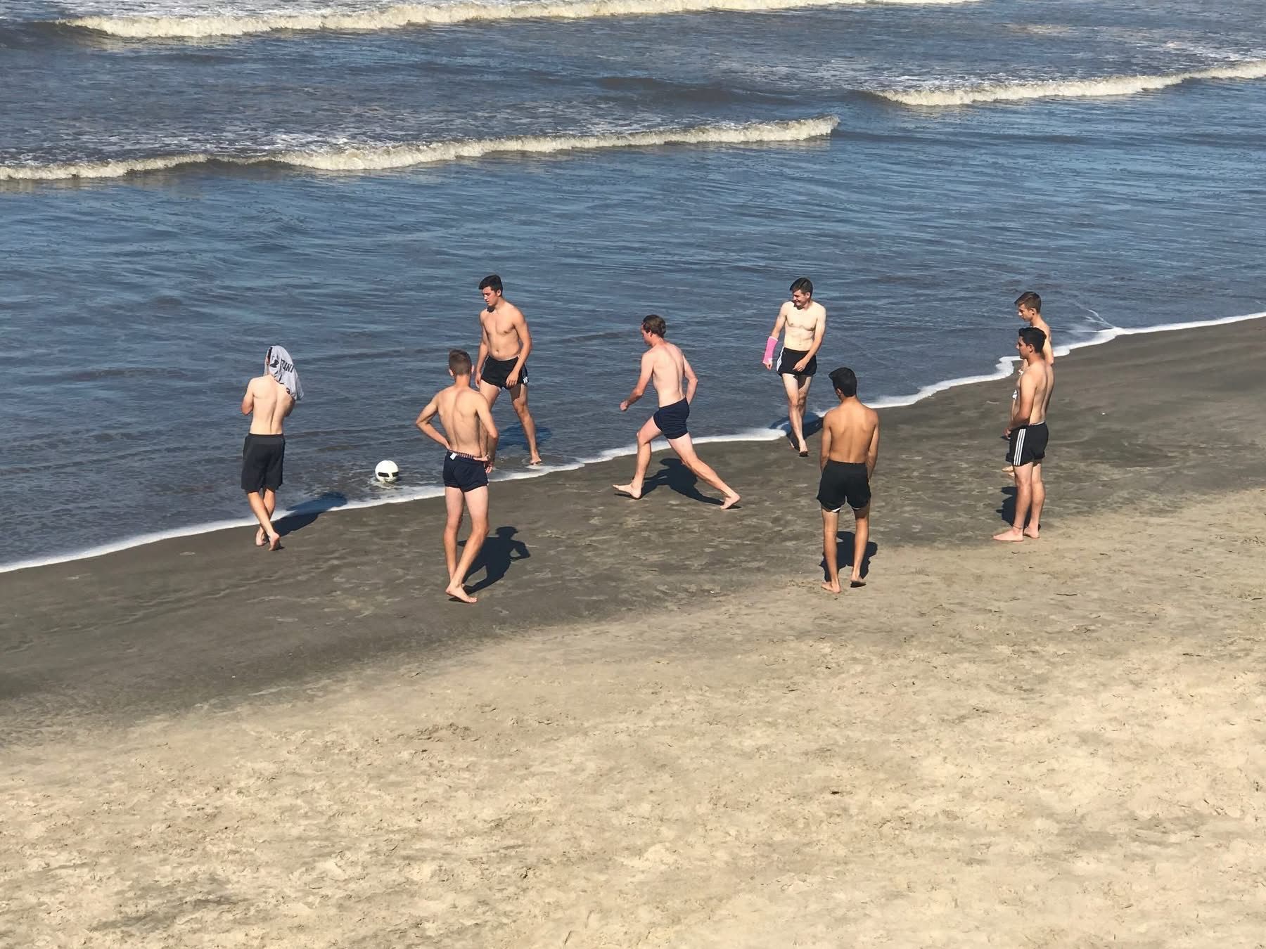 Men playing soccer on a sandy beach near the ocean; one kicking the ball.
