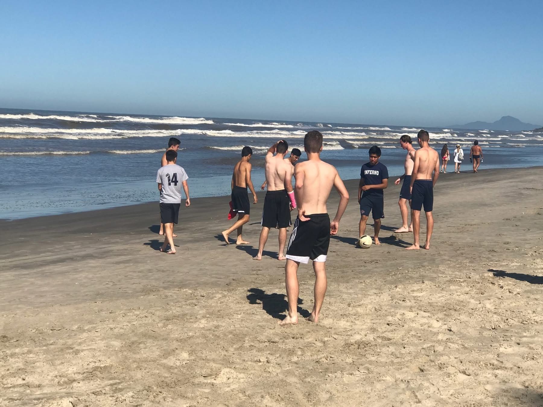 People playing soccer on a sandy beach near the ocean under a clear sky.