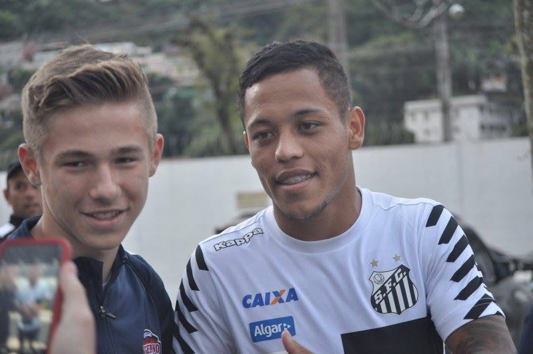 Soccer player with fan, smiling, posing for a photo outdoors. Player wears white and black jersey. Fan holds phone.