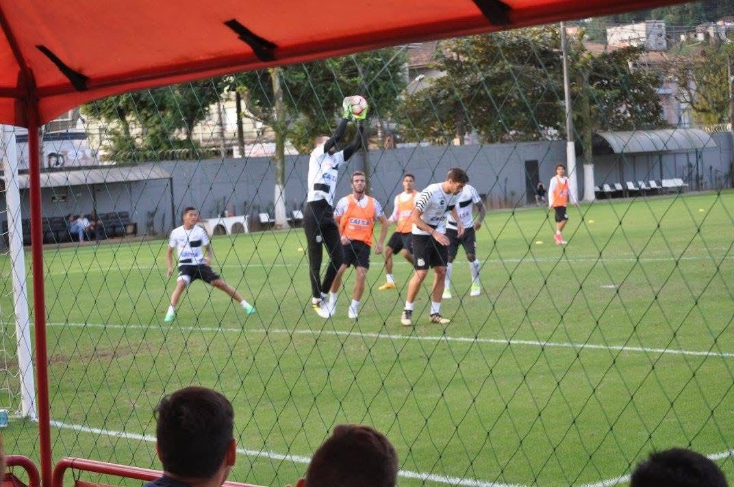 Soccer players practice on a green field. A goalkeeper jumps for the ball while others watch. A red awning frames the view.