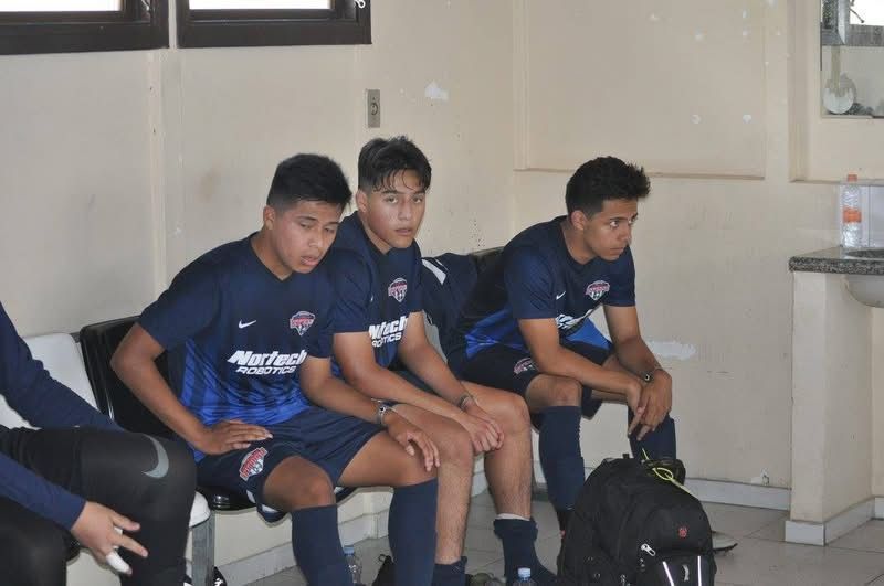 Three soccer players in navy blue uniforms sit on a bench indoors.