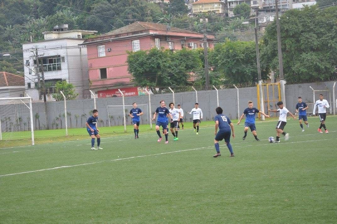 Soccer game on green field, players in blue and white uniforms, buildings in background.