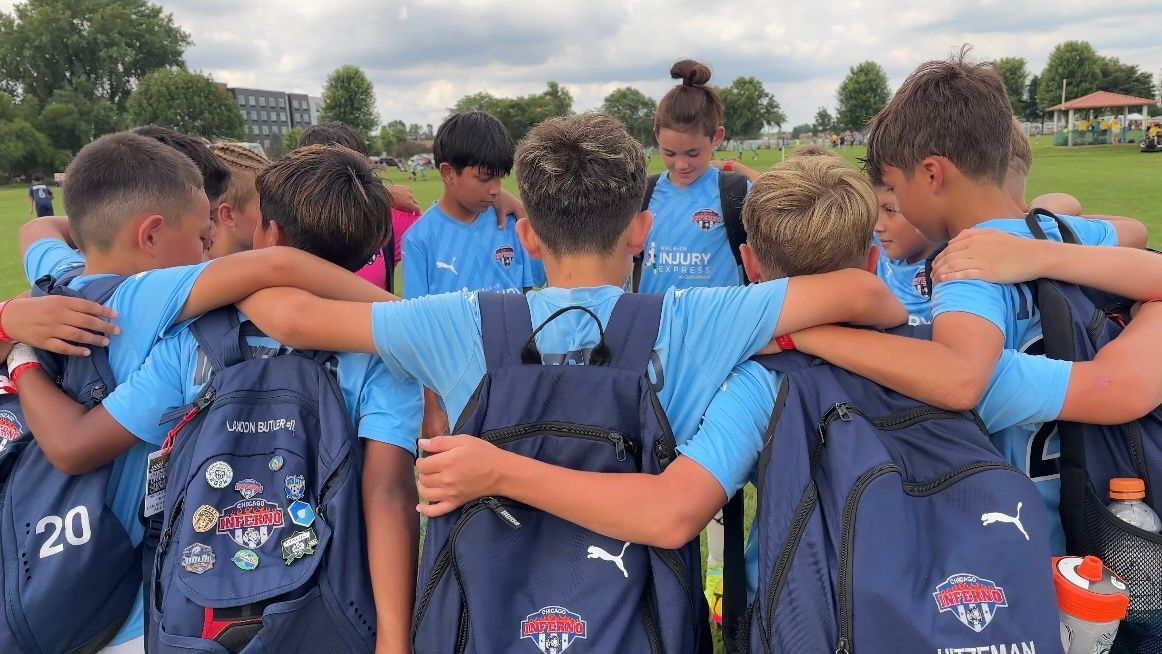 Soccer team huddles together, arms around each other. Wearing light blue jerseys and backpacks, on a field.