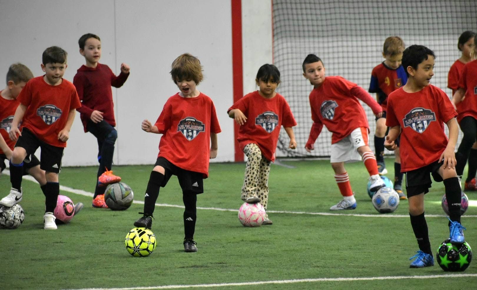 Kids in red shirts practice soccer indoors on green turf, dribbling balls.