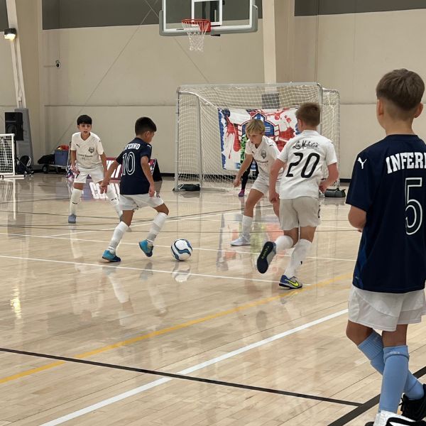 Indoor youth soccer game: boys in white and navy jerseys playing on a wooden court. One player dribbles the ball.
