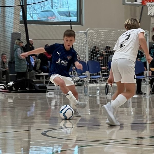 Soccer players in action indoors; one dribbles a ball, the other defends.