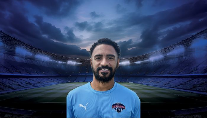 Coach Tayrones Oliveira in light blue jacket stands in front of a soccer stadium backdrop.