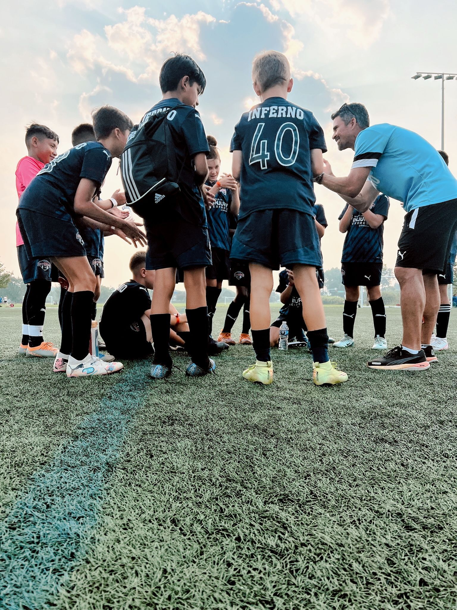 Youth soccer team in dark blue uniforms huddles with coach on a green field under a clear sky.