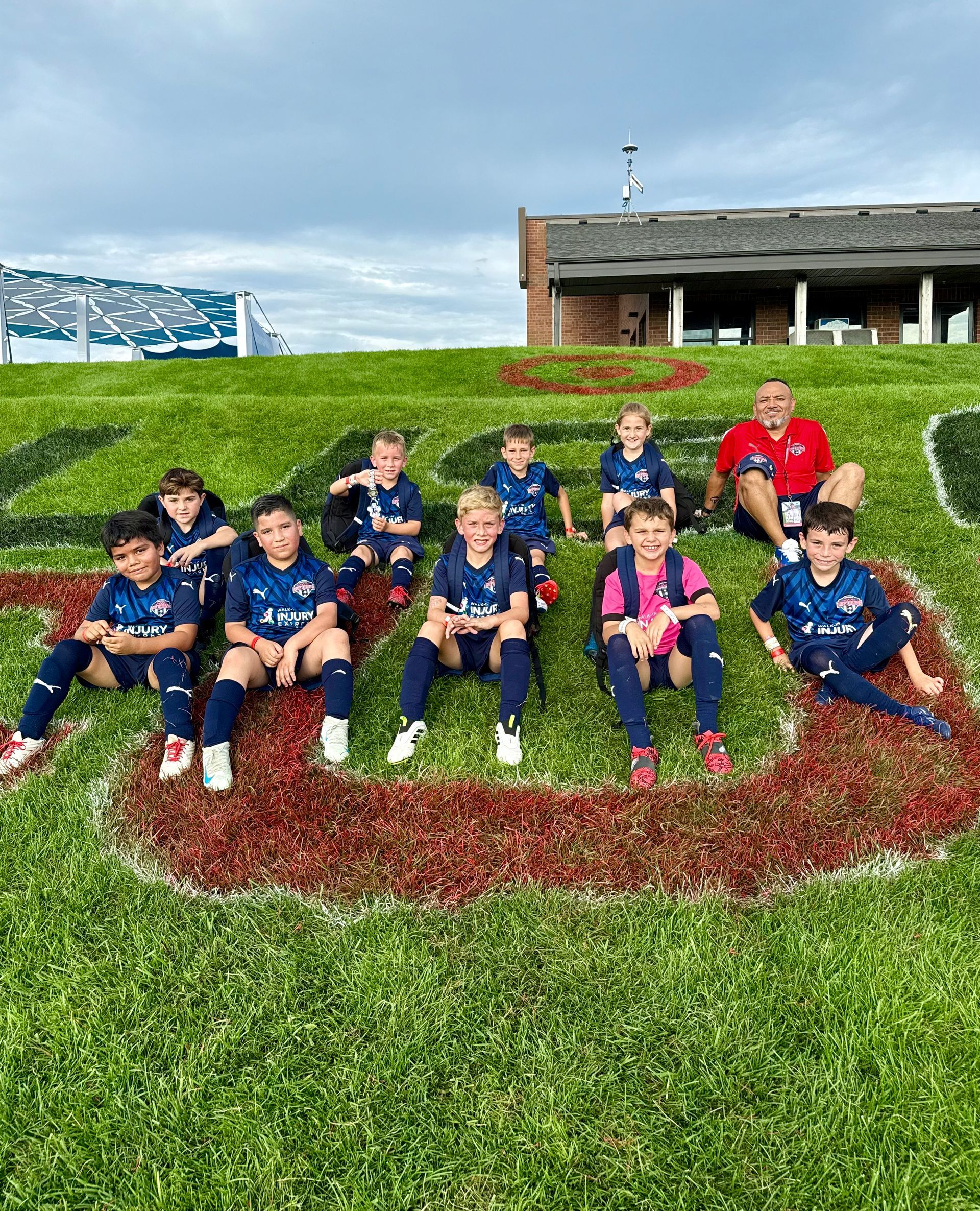 Youth soccer team poses on a grassy hill; players wear navy and pink jerseys in front of a building.