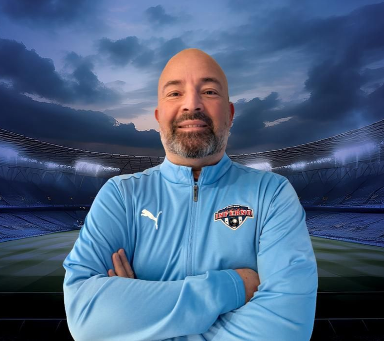 Coach Pierre Garcia  with a bald head and beard wearing a light blue jacket with arms crossed, in a soccer stadium setting.