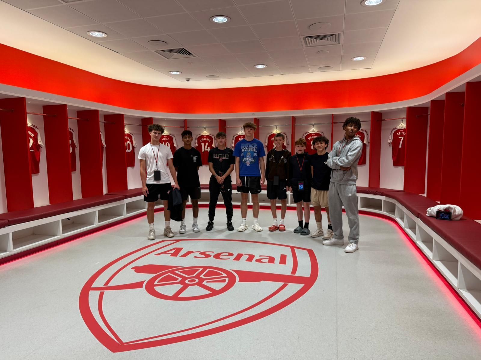 Group of people in an Arsenal locker room. The team emblem is on the floor.
