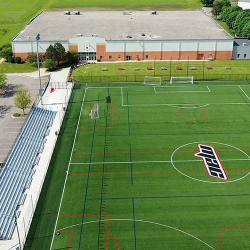 Soccer field with building in background; green turf, bleachers, and MAC logo.
